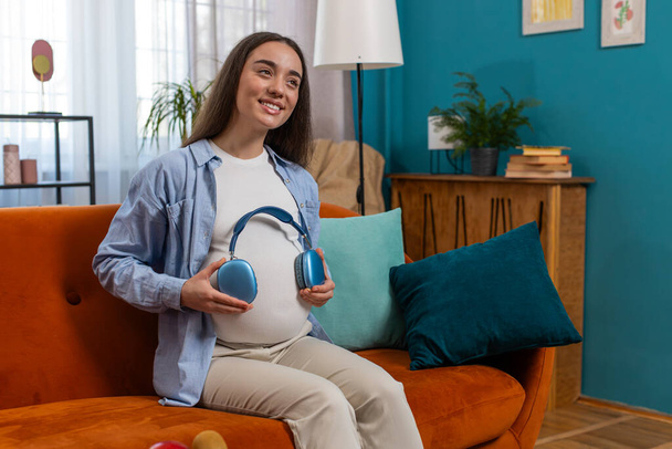 Щаслива вагітна жінка сидить на дивані і кладе бездротові навушники на животик з дитиною, слухаючи улюблену музику вдома. Технології, майбутнє матері та концепція вітчизняної діяльності - Фото, зображення