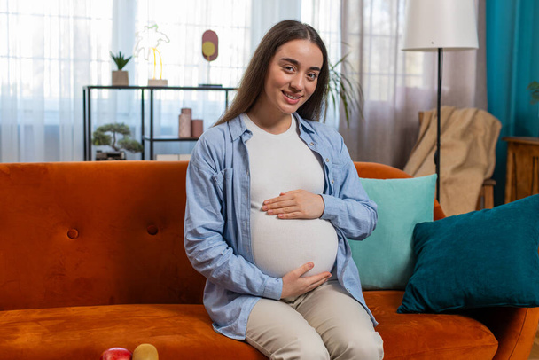 Portret van een glimlachende toekomstige moeder die op de bank zit en zwangere buik aanraakt in de woonkamer thuis. Gelukkig zwanger Kaukasische dame ontspannen op bank kijken naar camera zit in de buurt van zacht speelgoed in appartement. - Foto, afbeelding