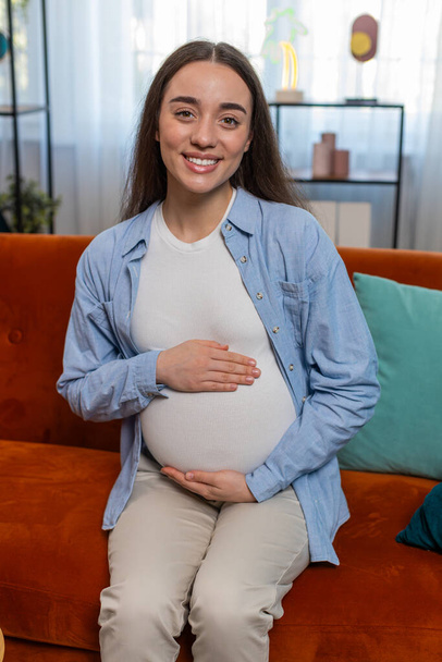 Портрет усміхненої майбутньої матері, яка сидить на дивані, торкаючись вагітного живота у вітальні вдома. Щаслива вагітна кавказька леді дівчина розслабляється на дивані, дивлячись на камеру сидить біля м'якої іграшки в квартирі - Фото, зображення