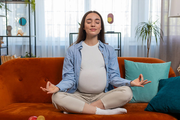 Calm pregnant charming woman sitting in lotus position while meditating on comfy sofa in living room at home. Concept of health care, pregnancy and breathing practice. Happy girl prepares to be mother - Photo, Image