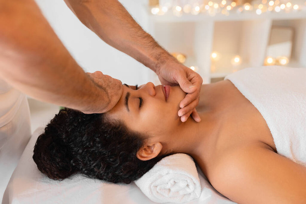 Um massagista qualificado oferece uma massagem facial relaxante para uma senhora afro-americana cliente em um ambiente tranquilo de spa com iluminação fraca e velas - Foto, Imagem