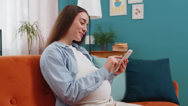 Smiling pregnant woman using smartphone text messaging social media chatting sitting on sofa in living room at home. Happy Caucasian lady future mother stroking belly making online purchases for baby - Footage, Video