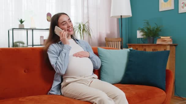 Uśmiechnięta ciężarna kobieta rozmawiająca na smartfonie siedząc na kanapie w salonie w domu. Szczęśliwa biała kobieta głaszcze brzuch trzymając komórkę w mieszkaniu. Pojęcie opieki prenatalnej i ciąży macierzyńskiej. - Materiał filmowy, wideo