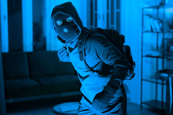Une silhouette sinistre dans un masque de ski avec un sac à dos se faufile dans une maison faiblement éclairée, suggérant vol, danger et crime. Le ton bleu ajoute à l'atmosphère étrange - Photo, image