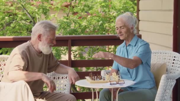 Medium shot of senior 70-plus Caucasian husband and wife sitting together in armchairs on terrace outdoors, playing chess, pointing to board, gesticulating and discussing game strategy - Footage, Video