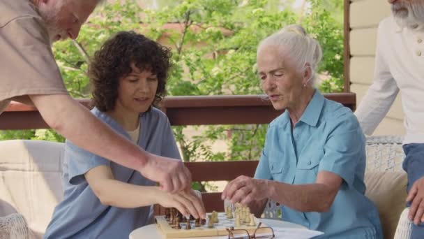 Imágenes medianas de la alegre enfermera joven en uniforme azul sentada en la mesa en la terraza con una mujer mayor caucásica, aprendiendo a jugar al ajedrez, recibiendo cheques y pidiendo ayuda a un residente masculino - Imágenes, Vídeo