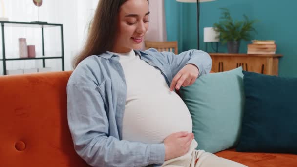 Счастливая молодая беременная женщина ходит пальцами по своему большому животу, сидя на диване в гостиной дома. Будущая мать играет с нерожденным ребенком в животе в современной квартире - Кадры, видео