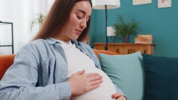 Zwangere vrouw die slaapliedje zingt en grote buik aanraakt zittend op de bank in de woonkamer thuis. Houden van toekomstige moeder dame praten met ongeboren kind baby besteedt vrije tijd op de bank. Nieuw gezinsconcept - Video