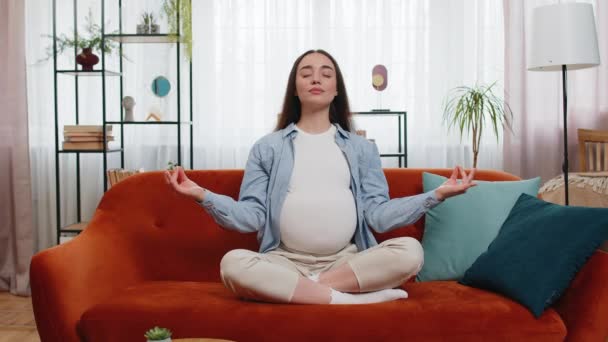 Calm pregnant charming woman sitting in lotus position while meditating on comfy sofa in living room at home. Concept of health care, pregnancy and breathing practice. Happy lady prepares to be mother - Záběry, video