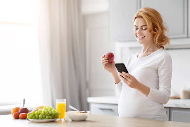 Mujer embarazada europea comprueba un teléfono móvil, con un enfoque en la nutrición, de pie en una cocina con frutas cerca - Foto, Imagen