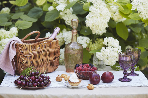 kosz piknikowy z kocem, butelka vintage, świeże owoce i fioletowe okulary, zestaw przeciwko krzewom hortensji - Zdjęcie, obraz