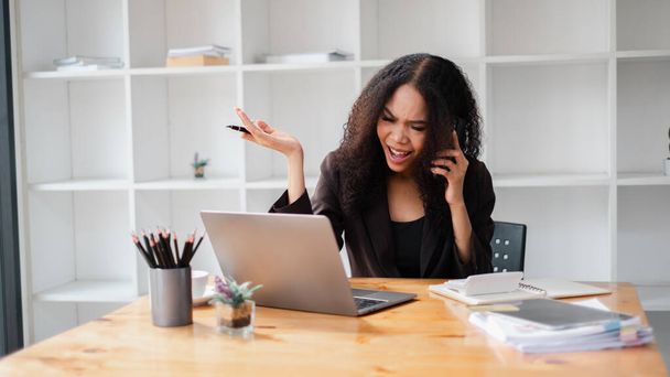 Femme d'affaires montrant de la frustration lors d'un appel téléphonique, avec un ordinateur portable et des documents sur son bureau. - Photo, image