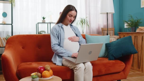 Happy pregnant woman touching belly while using laptop computer sitting on sofa in living room at home. Future Caucasian mother communicating online browsing internet social media. Pregnancy and job. - Footage, Video