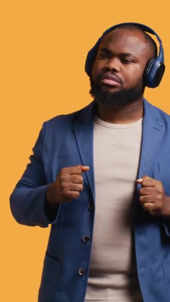 Függőleges videó Boldog afro-amerikai személy szórakozik, elektronikus dalokat hallgat fejhallgatón keresztül. Vidám BIPOC úgy tesz, mintha DJ, élvezi a tánc zene, stúdió háttér, kamera B - Felvétel, videó
