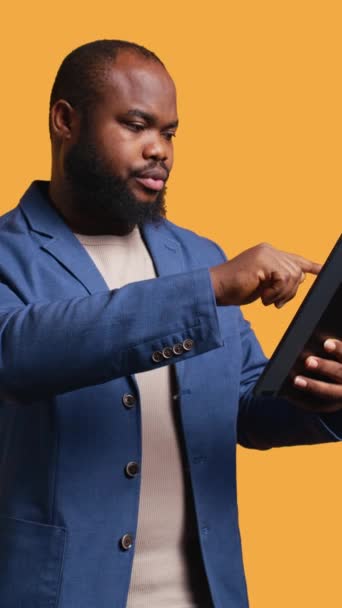 Vertical video Man happily scrolling on tablet touchscreen to check social media feed. African american person browsing internet websites, entertained by content, studio background, camera B - Footage, Video