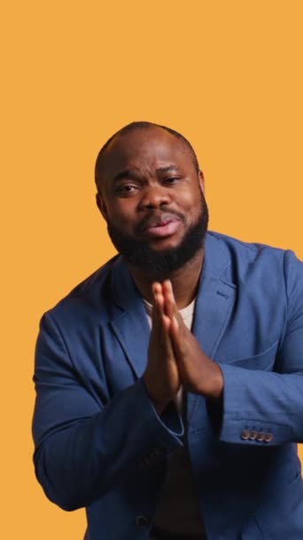 Verticale video Afro-Amerikaanse man die handen bij elkaar steekt in smekend gebaar, wensend, geïsoleerd over gele studio achtergrond. BIPOC persoon vraagt om iets, bidden en hopen, camera B - Video
