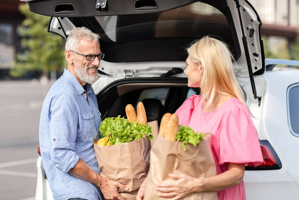 Una pareja de ancianos comparte un momento junto a su coche, con la compra en la mano, mostrando su duradera vida matrimonial y compañía - Foto, Imagen
