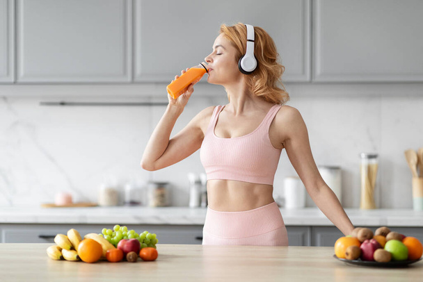 In een moderne keuken, een sportieve Europese dame snacken op een wortel vertegenwoordigt haar bewuste voeding keuzes na de training - Foto, afbeelding
