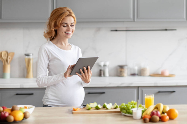 Μια ευρωπαϊκή έγκυος κυρία στέκεται σε μια σύγχρονη κουζίνα περιήγηση σε ένα tablet, που περιβάλλεται από φρέσκια διατροφή - Φωτογραφία, εικόνα