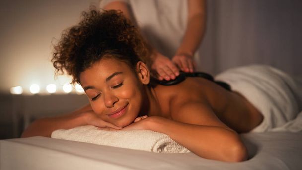 Capturada en un ambiente suavemente iluminado, una dama afroamericana se muestra tranquila durante una sesión de masaje de spa centrada en la relajación y el bienestar. - Foto, imagen