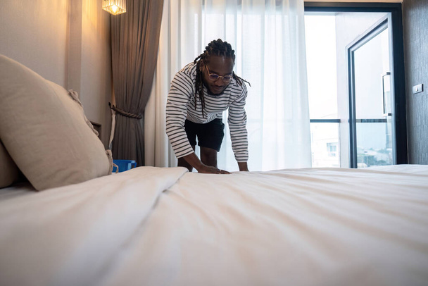 Młody Afroamerykanin ścieli łóżko. szczęśliwy czarny człowiek zmiana pościel w domu stojąc w sypialni. - Zdjęcie, obraz