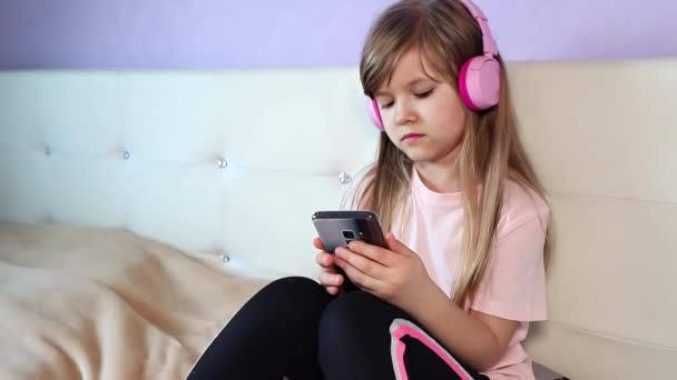 Linda niña preadolescente sonriente en camiseta rosa con auriculares rosas escuchar música y un poco de baile por la cabeza utilizar un teléfono inteligente, sentado en una cama en casa, cámara lenta. Imágenes de alta calidad 4k - Metraje, vídeo