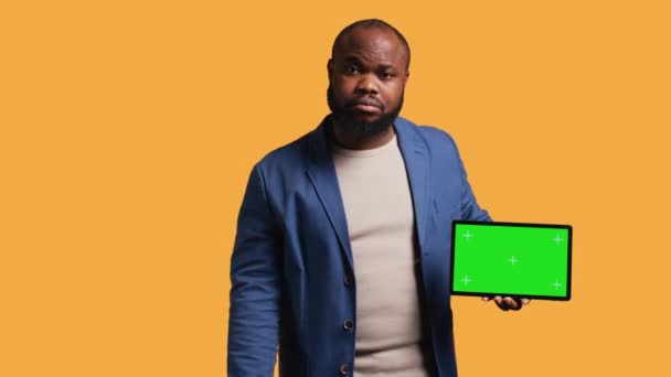 Upset african american man shows thumbs down sign gesturing holding isolated screen tablet, disapproving. BIPOC person does rejection hand gesture regarding mockup device, studio background, camera B - Footage, Video