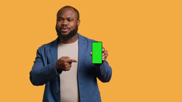 Usmívající se afroameričan prezentující mobilní telefon se zeleným displejem, izolovaný na pozadí studia. Veselý BIPOC osoba vytváří propagaci s prázdnou kopií prostor mokup telefon, fotoaparát A - Záběry, video