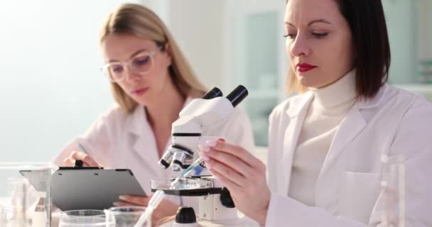 Kolega obserwuje próbki pod mikroskopem w pobliżu kobiety notuje szczegóły. Kobiety świadczą o wspólnym charakterze badań naukowych w zwolnionym tempie - Materiał filmowy, wideo