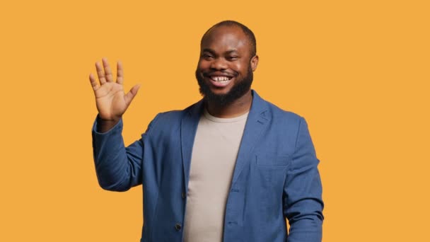 Африканский американец с радостью делает приветственный жест рукой. Портрет счастливого человека BIPOC, поднимающего руку, чтобы поприветствовать кого-то, жестикулирующий, изолированный на жёлтом фоне студии, камера А - Кадры, видео