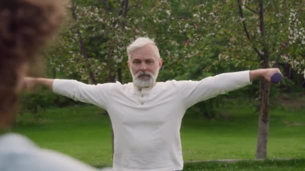 Média sobre ombro tiro de idoso caucasiano com cabelos grisalhos e barba exercitando-se no jardim de casa convalescente, fazendo levantamento lateral com halteres, durante a fisioterapia - Filmagem, Vídeo