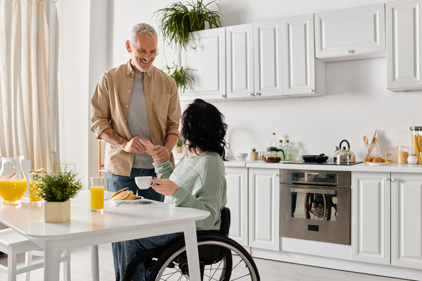 Ένας σύζυγος στέκεται δίπλα στην ανάπηρη γυναίκα του σε μια αναπηρική καρέκλα στην άνετη κουζίνα του σπιτιού τους, μοιράζονται μια στιγμή συντροφικότητας. - Φωτογραφία, εικόνα