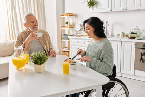 Μια ανάπηρος γυναίκα σε αναπηρική καρέκλα και ο σύζυγός της μοιράζονται μια ζεστή στιγμή στο τραπέζι της κουζίνας στο σπίτι τους. - Φωτογραφία, εικόνα