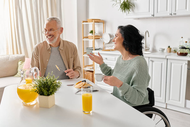 Egy férfi és egy nő, a tolószékes nő, beszélgetnek egy asztalnál egy hangulatos konyhában.. - Fotó, kép