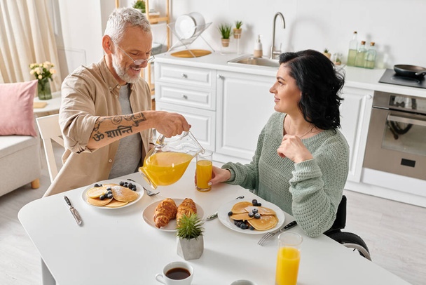 Une femme handicapée en fauteuil roulant et son mari prennent le petit déjeuner ensemble à une table de cuisine dans leur confortable maison. - Photo, image