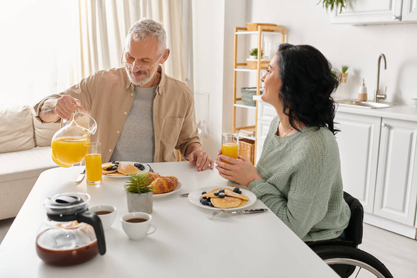 Μια ανάπηρος γυναίκα σε αναπηρική καρέκλα και ο σύζυγός της απολαμβάνουν ένα ήρεμο πρωινό γεύμα μαζί στο τραπέζι της κουζίνας τους. - Φωτογραφία, εικόνα