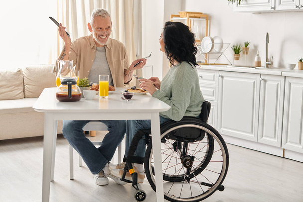 Ένας άνδρας σε αναπηρική καρέκλα και μια γυναίκα απολαμβάνουν ένα γεύμα μαζί στην κουζίνα τους στο σπίτι. - Φωτογραφία, εικόνα