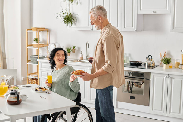 Eine Frau im Rollstuhl hält einen Teller Croissant in der Hand, während sie mit ihrem Partner in einer gemütlichen Küche zu Hause interagiert. - Foto, Bild