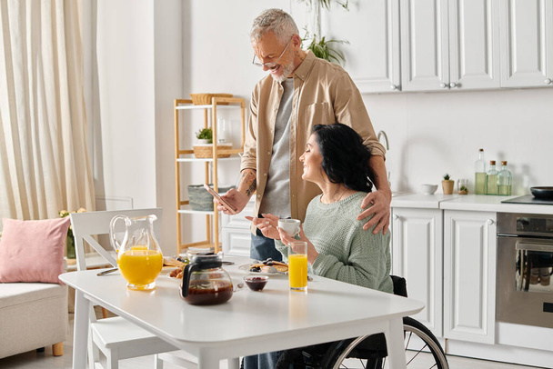 Ένας σύζυγος στέκεται δίπλα στην ανάπηρη γυναίκα του σε αναπηρική καρέκλα, προσφέροντας υποστήριξη και συντροφιά στην κουζίνα του σπιτιού τους. - Φωτογραφία, εικόνα