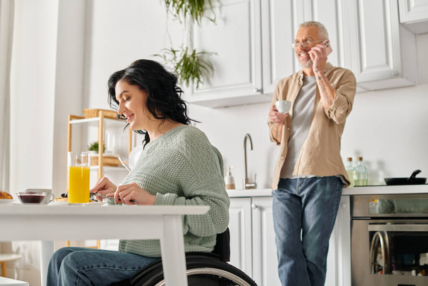 Μια ανάπηρος γυναίκα σε αναπηρική καρέκλα και ο σύζυγός της μαζί στην κουζίνα του σπιτιού τους. - Φωτογραφία, εικόνα