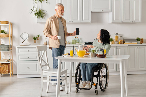 Una donna disabile in sedia a rotelle che fa conversazione con il marito in una cucina accogliente a casa. - Foto, immagini