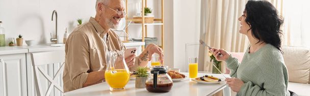 Ένας άντρας και μια ανάπηρη γυναίκα σε αναπηρικό καροτσάκι μοιράζονται ένα ήρεμο πρωινό γεύμα σε ένα άνετο τραπέζι κουζίνας στο σπίτι τους.. - Φωτογραφία, εικόνα