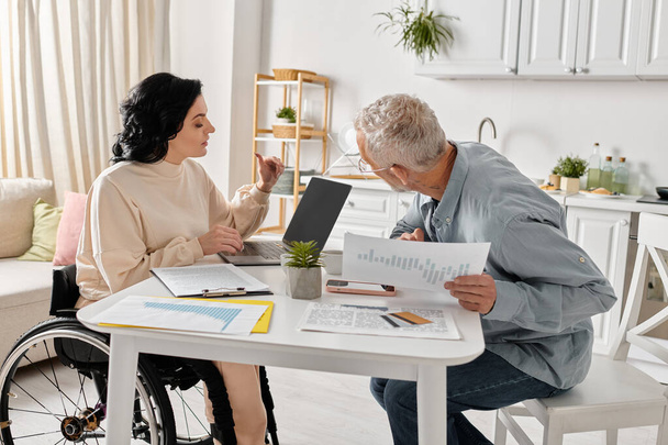 Чоловік сидить за столом поруч зі своєю дружиною в інвалідному візку на їхній кухні вдома, дивлячись на ноутбук - Фото, зображення