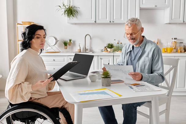 Ein Mann sitzt mit seiner behinderten Frau im Rollstuhl an einem Tisch, beide konzentriert auf einem Laptop-Bildschirm in ihrer gemütlichen Küche zu Hause.. - Foto, Bild