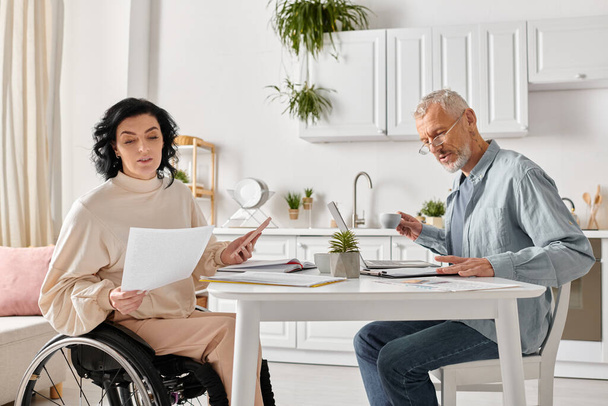 Una mujer en silla de ruedas sosteniendo el teléfono y un hombre con un portátil, compartiendo un momento de unión en su cocina en casa. - Foto, imagen