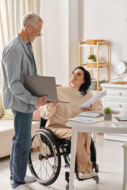 Μια ανάπηρος γυναίκα σε αναπηρική καρέκλα και ο σύζυγός της συνεργάζονται σε ένα φορητό υπολογιστή στην κουζίνα τους στο σπίτι. - Φωτογραφία, εικόνα