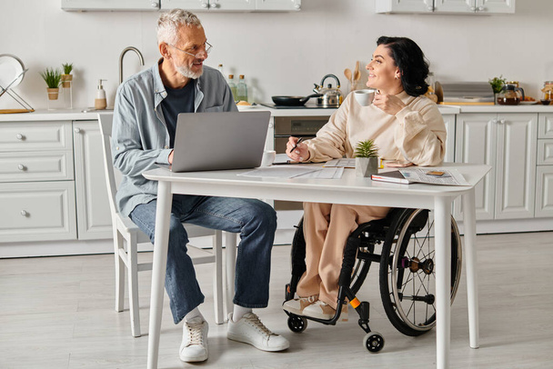 Un marido y su esposa discapacitada se sientan en una mesa de la cocina trabajando juntos en un portátil. - Foto, imagen