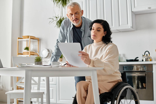 Ένας άνδρας σε αναπηρική καρέκλα και μια γυναίκα εξετάζουν ένα έγγραφο σε ένα άνετο περιβάλλον κουζίνας στο σπίτι. - Φωτογραφία, εικόνα