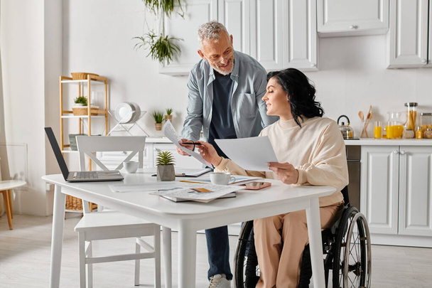 Ένας άντρας και μια γυναίκα σε αναπηρικά καροτσάκια ελέγχουν προσεκτικά ένα κομμάτι χαρτί στην κουζίνα τους στο σπίτι.. - Φωτογραφία, εικόνα