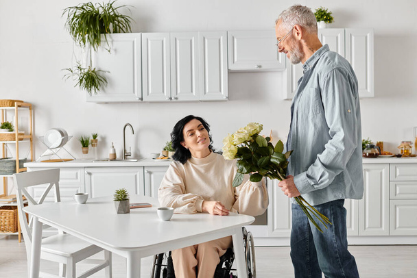 Un homme se tient tendrement à côté de sa femme dans un fauteuil roulant, partageant un moment de connexion et de soutien dans leur cuisine à la maison. - Photo, image